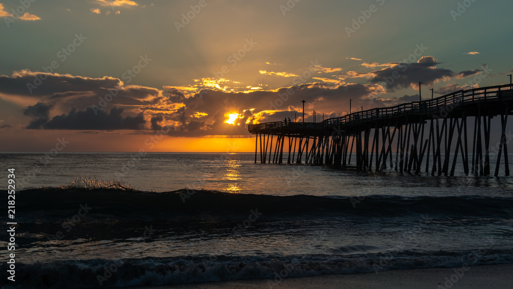 A golden sunrise over the Atlantic Ocean in North Carolina. Small waves break on the shore. A fishing pier extends far out into the sea.