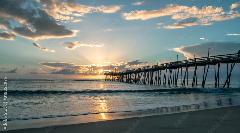 Sunrise behind light clouds lights the sky as the ocean reflects the morning sun. A long fishing pier and flat sea.