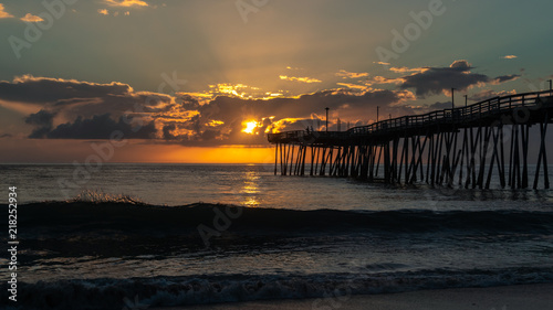 A golden sunrise over the Atlantic Ocean in North Carolina. Small waves break on the shore. A fishing pier extends far out into the sea.