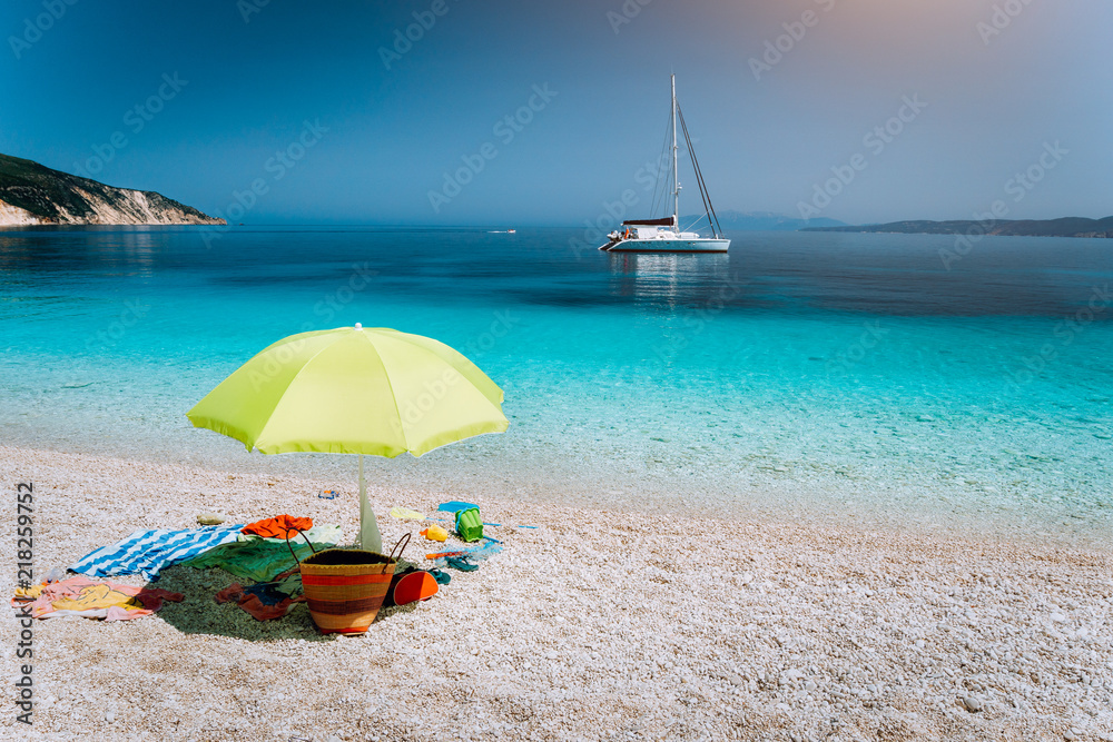 Idyllic white beach with umbrella on lazy summer day. Sailing boat at anchor in calm crystal clear blue sea water