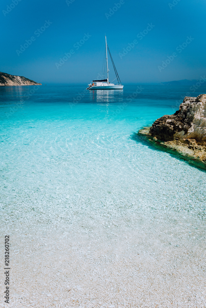 Fototapeta premium Azure blue lagoon with sailing catamaran yacht boat at anchor. Pure white pebble beach, some rocks in the sea
