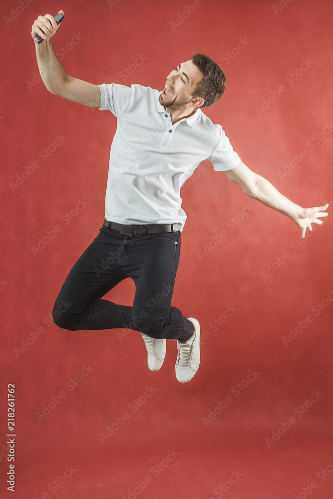 Photo of excited bearded man jumping over red background wall using mobile phone make selfie.