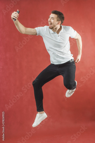 Photo of excited bearded man jumping over red background wall using mobile phone make selfie. © Виталий Давыдов