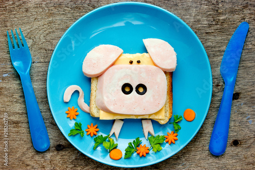 Fun food art idea for kids breakfast - funny pig sandwich with cheese, ham and olives on blue plate top view