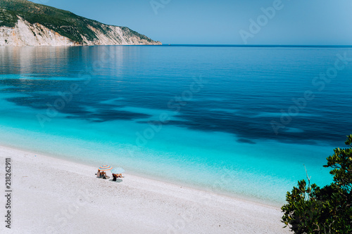 Fototapeta Naklejka Na Ścianę i Meble -  Fteri beach, Kefalonia, Greece. Lonely unrecognizable tourist couple hiding from sun umbrella chill relax near clear blue emerald turquoise sea water lagoon