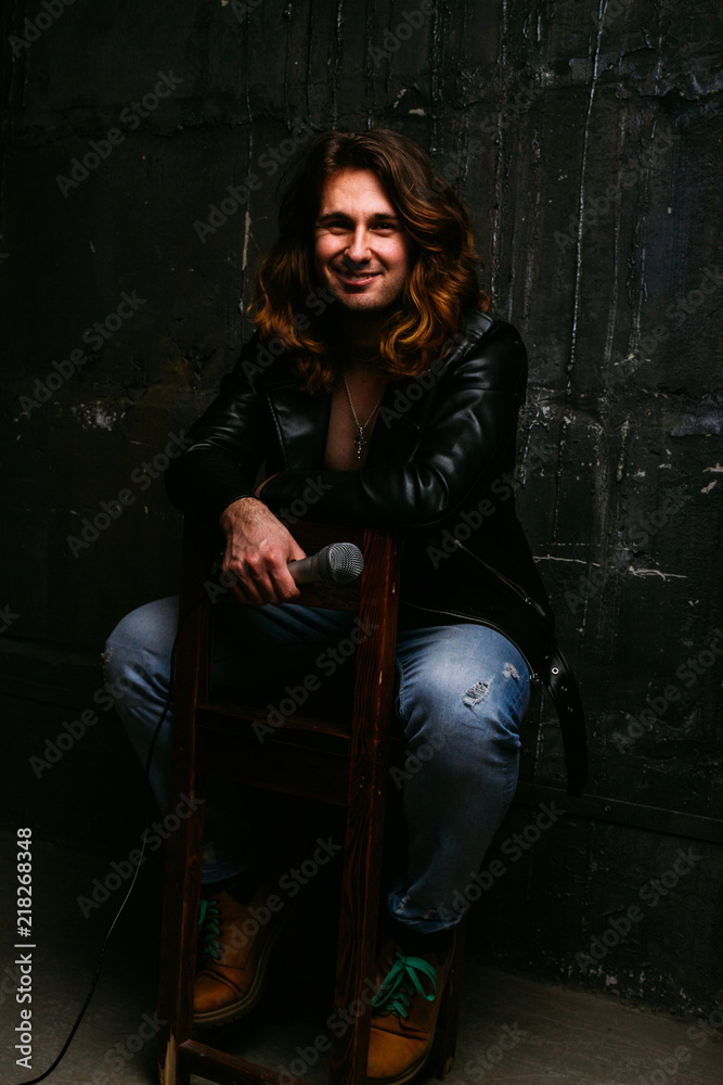 a portrait of a brutal, handsome man, a singer who sits on a chair on a dark background, in a black leather kurt on his naked body and holds a microphone in his hands. presenter, singer at the concert a portrait of a brutal, handsome man, a singer who sits on a chair on a dark background, in a black leather kurt on his naked body and holds a microphone in his hands. presenter, singer at the concert