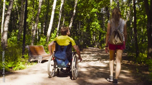 Dolly shot. Young disable man in a wheelchair on a walk in the park with his wife. Man riding in wheelchair himself. Rear view.