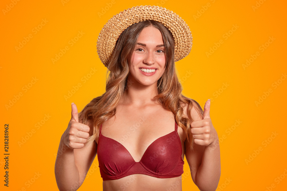 Horizontal shot of cheerful European female with broad smile and tanned skin makes okay gesture, wears red bikini and straw hat, shows her approval, likes resort place, isolated over orange wall