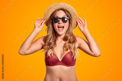 Pretty overjoyed female tourist keeps hands on summer hat, exclaims with happiness, has slim body, wears bikini, wears for beach season, isolated over orange bright background. Rest concept.