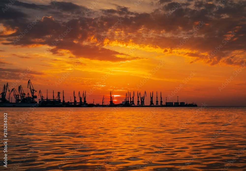 Beautiful landscape with seaport, fiery sunset sky and sea. Harbor on the coast during sunrise. Cranes silhouettes against fiery, orange and red sky.