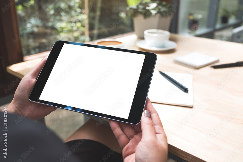 Mockup image of a woman's hands holding black tablet pc with white blank screen with notebook and coffee cup on table