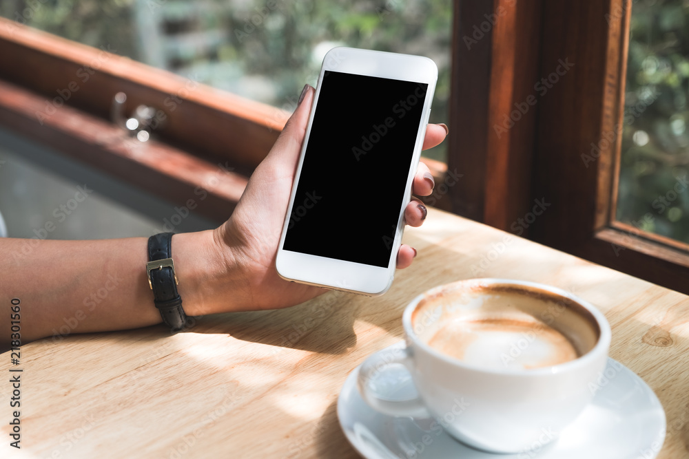 Mockup image of a hand holding and showing white mobile phone with blank black desktop screen with coffee cup on wooden table in cafe