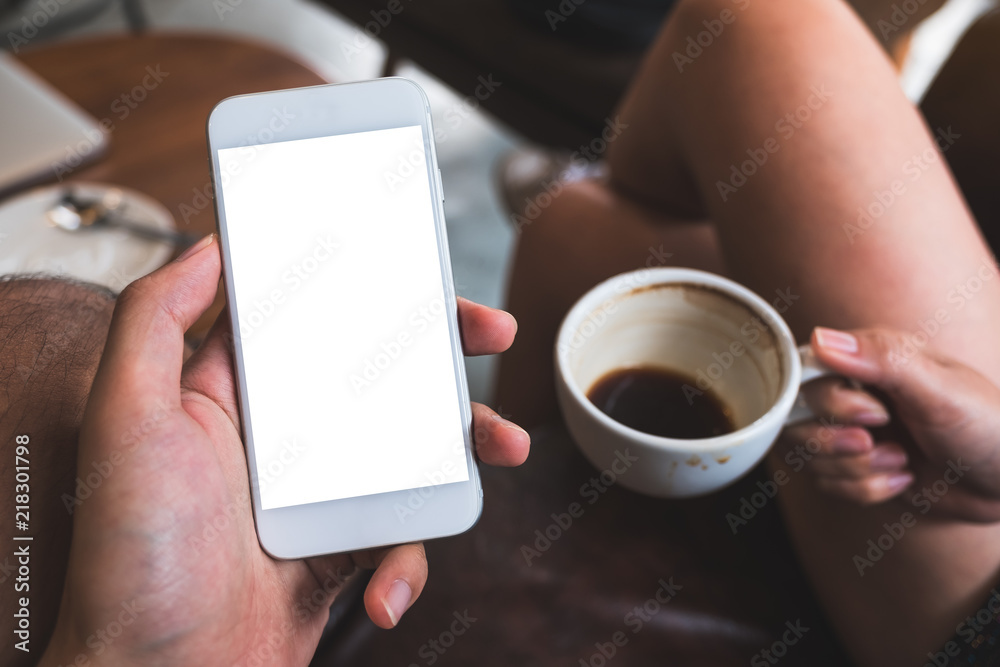 Mockup image of a hand holding white mobile phone with blank desktop screen with a woman holding black coffee cup while sitting on sofa with friend in cafe