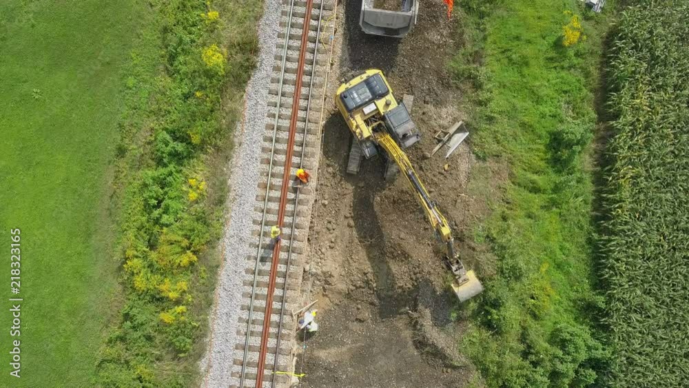 Aerial: Flying over excavator working at railway reconstruction site. Filming rails renovation at railway beside beautiful river on sunny day.