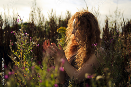 Woman with beautiful cascading long red hair standing amongst wild flowers highlighted by the sun 
