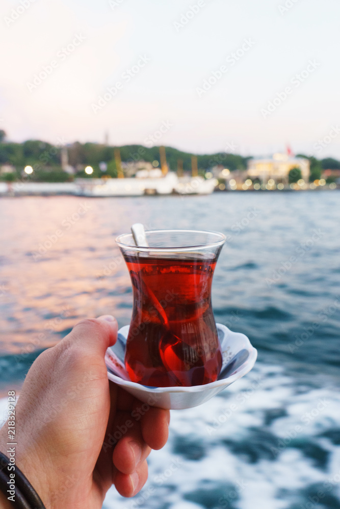 Traditional black Turkish tea in a glass on a plate in the hands with a view of the Bosporus and Istanbul. Traditional black Turkish tea in a glass on a plate in the hands with a view of the Bosporus and Istanbul.