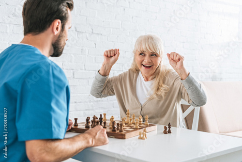 triumphing senior woman celebrating victory and looking at young social worker after chess game