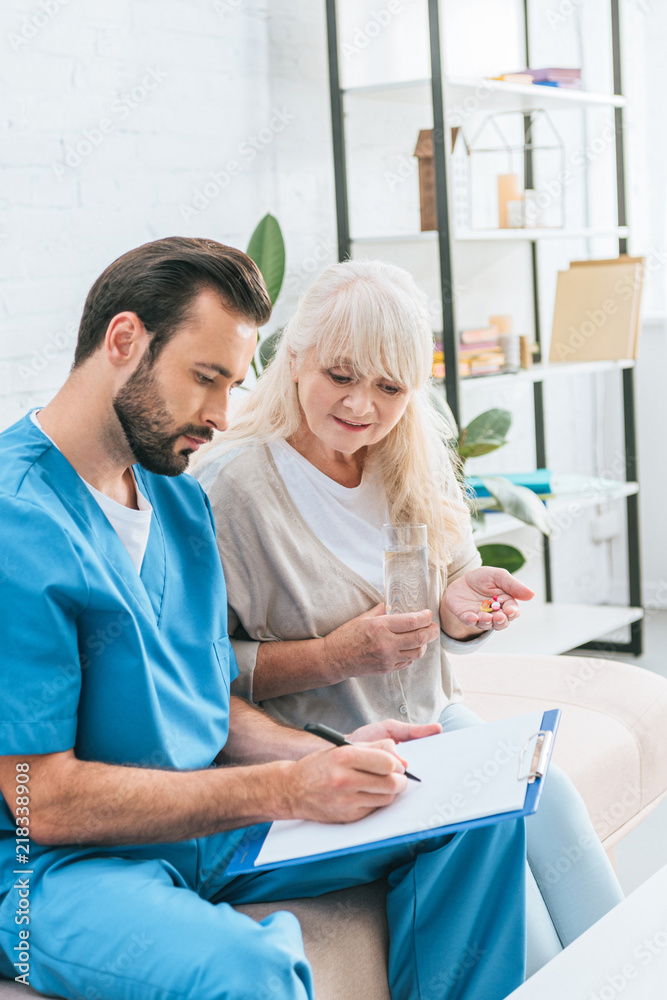Fototapeta premium senior woman taking pills and looking at male nurse writing on clipboard