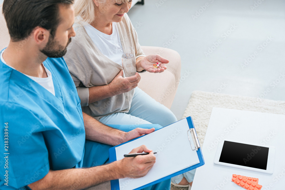 Obraz premium high angle view of male nurse writing on clipboard and senior woman taking pills