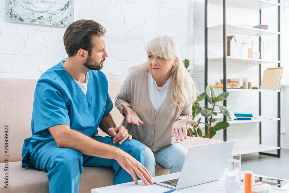 Fototapeta premium senior woman and young caregiver looking at each other while using laptop together