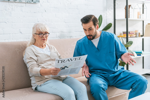 shocked male nurse looking at senior woman in eyeglasses reading travel newspaper photo