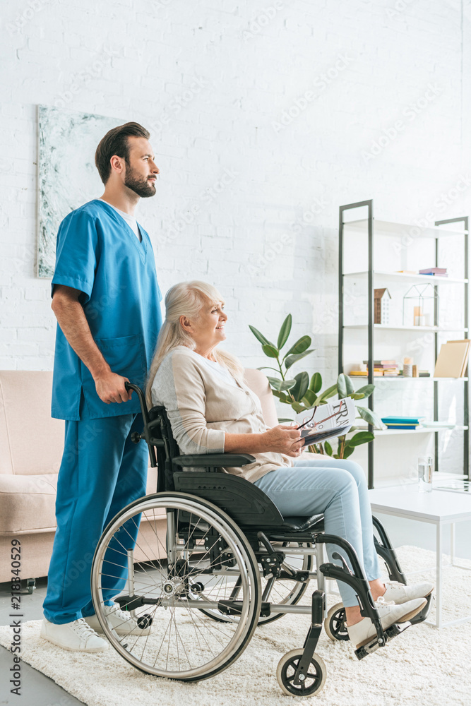 side view of social worker pushing wheelchair with senior woman holding newspaper and eyeglasses side view of social worker pushing wheelchair with senior woman holding newspaper and eyeglasses