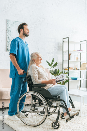 side view of social worker pushing wheelchair with senior woman holding newspaper and eyeglasses