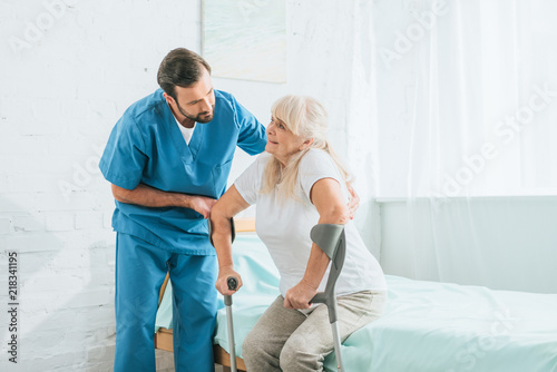 Obraz na płótnie male social worker helping sick senior woman with crutches