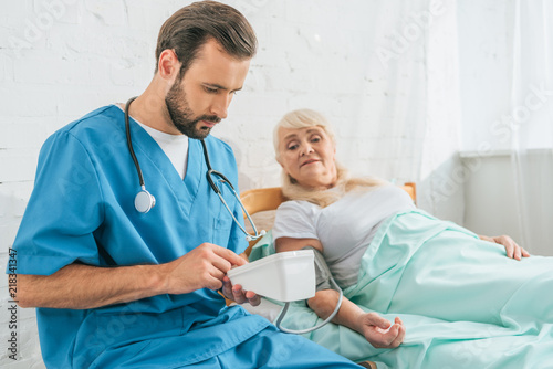 young male nurse with stethoscope holding blood pressure monitor while measuring blood pressure to senior woman lying in bed