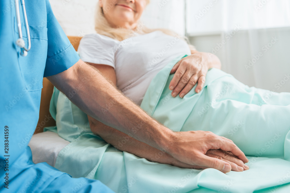 Obraz premium cropped shot of male nurse holding hand of sick senior woman lying in hospital bed