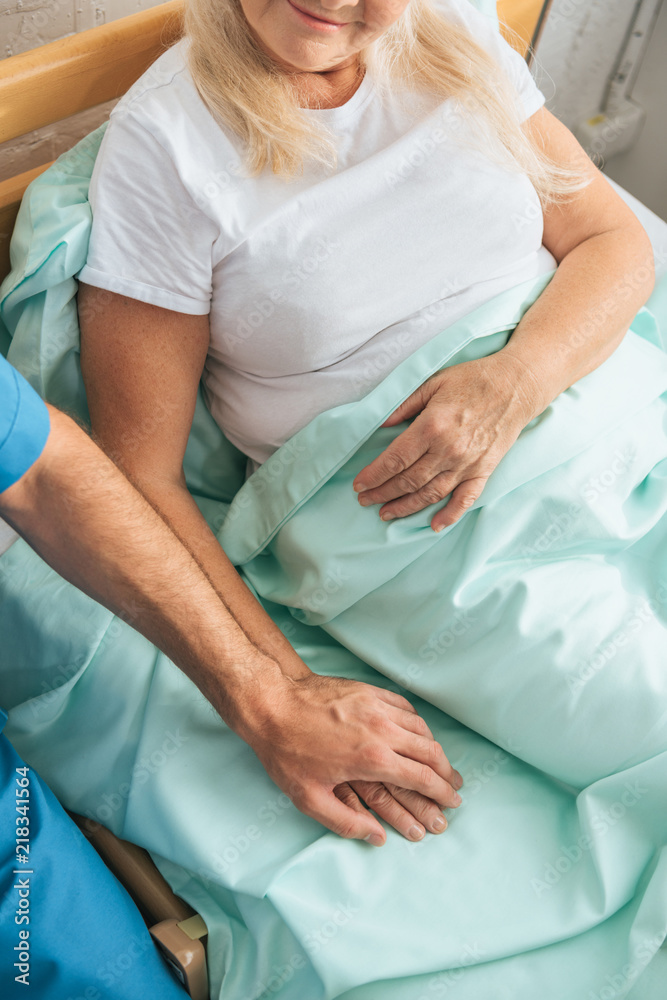 partial view of male nurse holding hand of sick senior woman lying in hospital bed