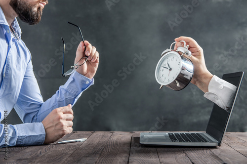 Businessman is sitting at the desk and is looking at the hand with clock coming out of the laptop. Metaphor of spending time at work