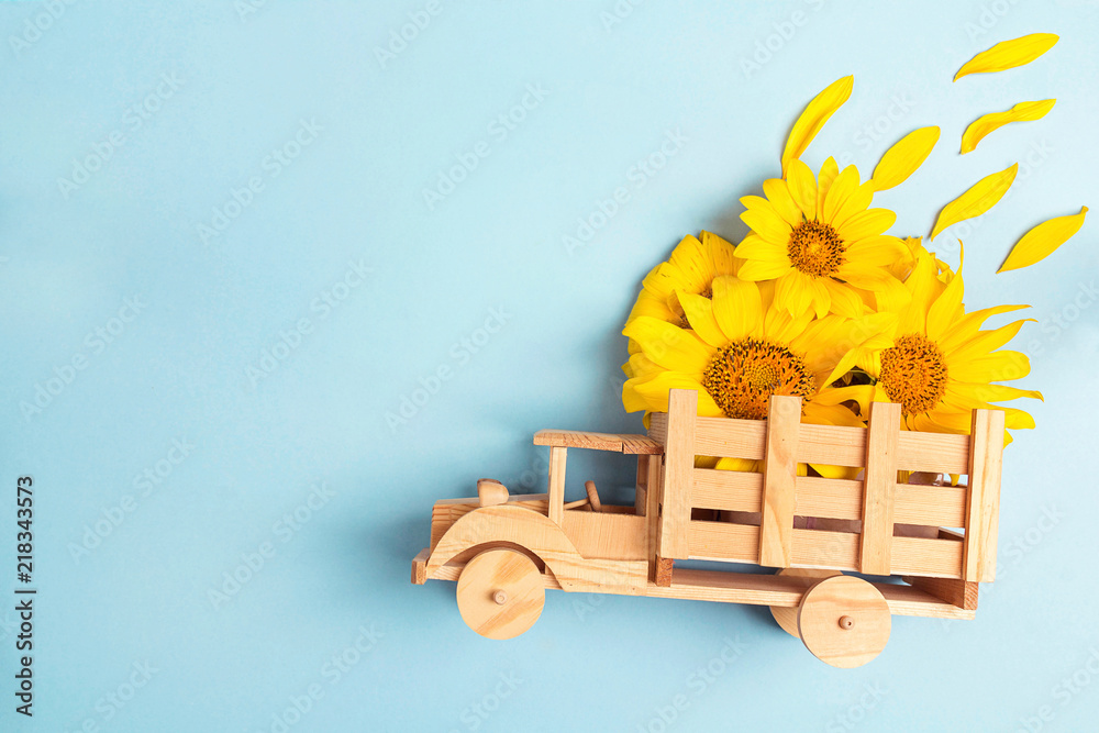 Fototapeta premium Drewniana zabawka ciężarówka z żółtymi słonecznikami z tyłu na niebieskim tle. Miejsce na tekst.