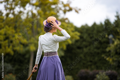 Beautiful girl in a vintage dress. Girl on a blurred background. A girl in a white blouse and a purple dress. A woman is dressed in retro style. The girl is straightening the hat on her head.