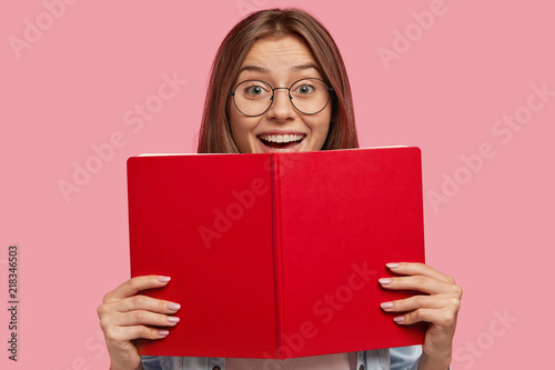 Happy European female student in spectacles, has positive expression, holds red book, rejoices successfully passed exam at university, isolated over pink background. People, learning, reading