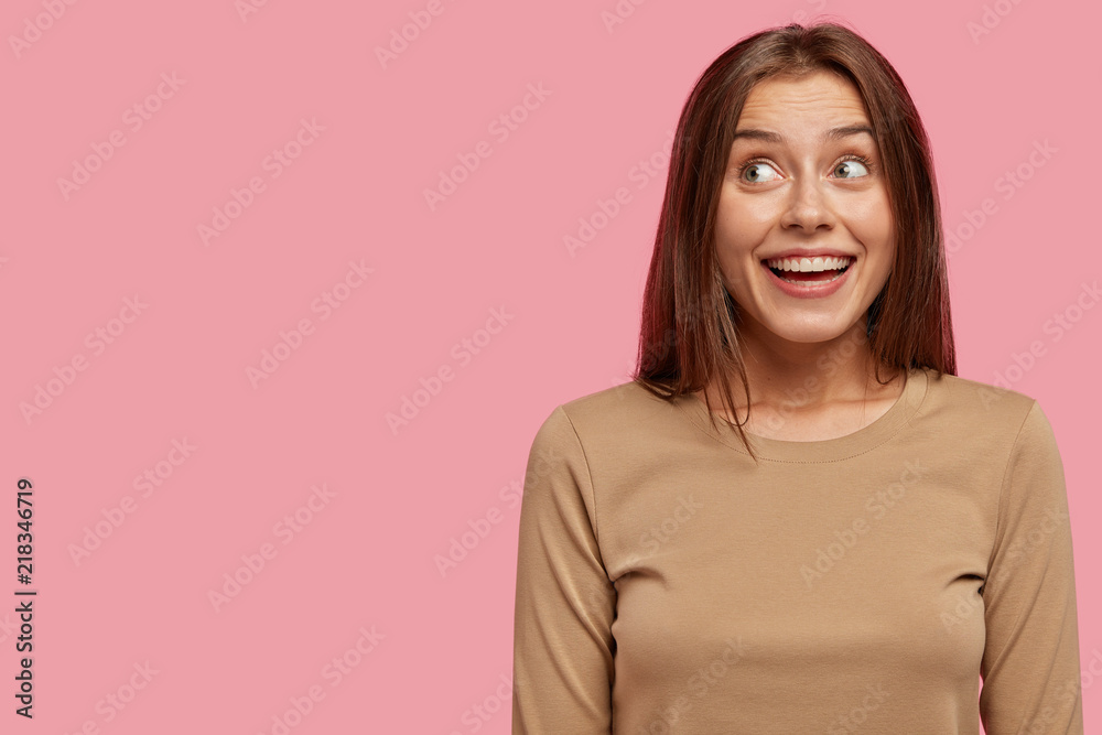 Studio shot of glad European young female has broad smile, looks positively and with dreamy expression aside, dressed in casual sweater, isolated over pink background with copy space on left Studio shot of glad European young female has broad smile, looks positively and with dreamy expression aside, dressed in casual sweater, isolated over pink background with copy space on left
