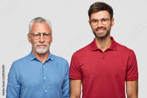 Mature grey haired man and his adult son stand against white background, have pleased expressions after meeting, wear round spectacles, being one friendly family. People and generation concept