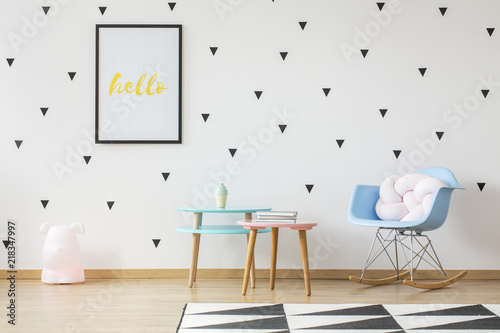 Fun wooden tables, light pink pillow in a baby blue rocking chair and a toy lamp in a cute nursery room interior with triangle stickers on a white wall © Photographee.eu