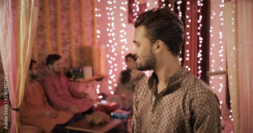 A handsome man in a traditional cloths smiles in interior house setup decorated with lights and flowers. A male wearing Kurta smiling during Diwali festival while group of friends talking in the back photo