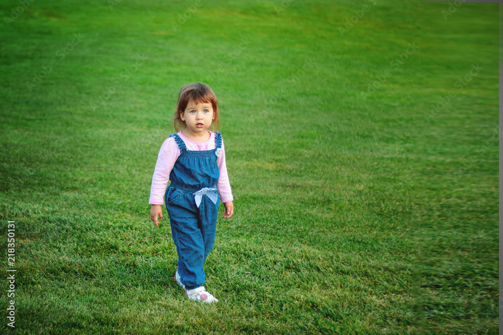 Little Caucasian girl with an amazed face stands on a green meadow in a blue overall, copyspase