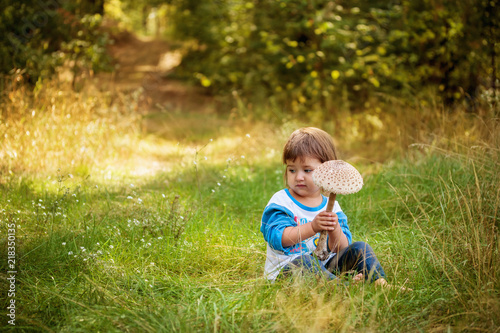 A smiling girl of two years is sitting on the grass and holding an edible mushroom