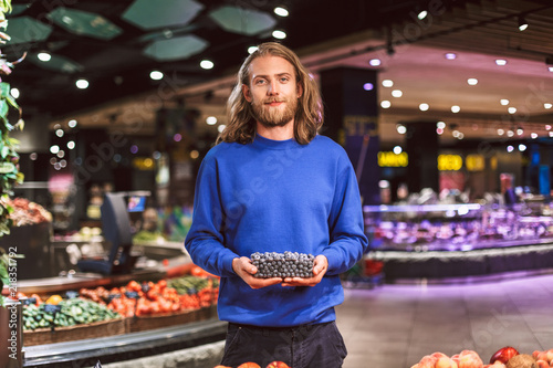 Young smiling man in dark blue sweater dreamily looking in camera while holding blueberries in hand in modern supermarket
