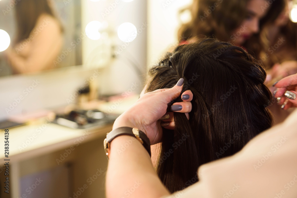 Fototapeta premium Beautiful girl with long hair, hairdresser weaves a French braid, in a beauty salon. Professional hair care and creating hairstyles.