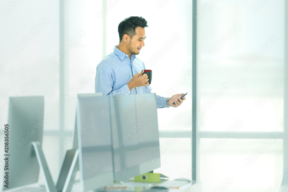 Young Asian businessman holding coffee cup and looking at smatphone smiling in calm the modern office