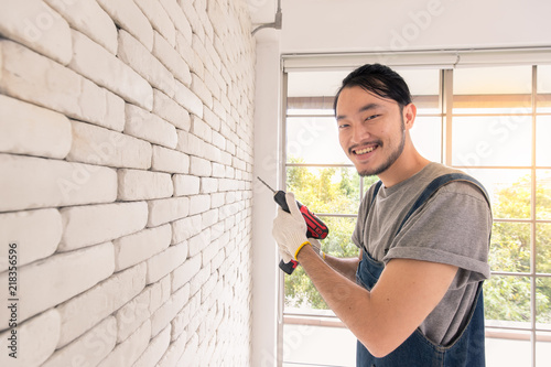 Young Asian man using electric drill on white brick wall in room, concept for home improvment, DIY work, do it youself photo