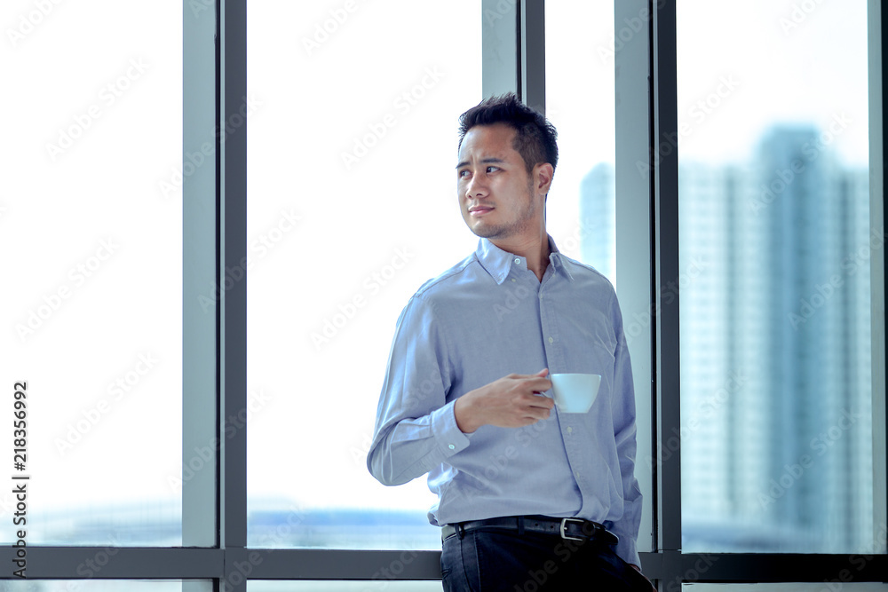 Fototapeta premium Young Asian businessman standing against window relaxed in his hands is cup of coffee look ahead.