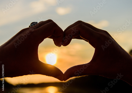 Sunset sunlight romantic atmosphere. Male hands in heart shape gesture symbol of love and romance. Heart gesture in front of sunset above river water surface, defocused. Top places for romantic date