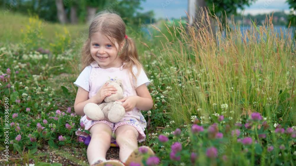 A little pretty girl plays with a plush rabbit, she sits in a meadow among a flowering clover. A little pretty girl plays with a plush rabbit, she sits in a meadow among a flowering clover.