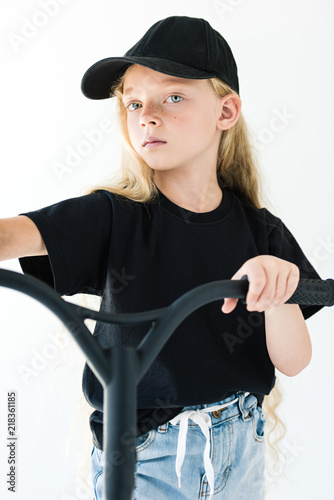adorable child in black t-shirt and cap riding scooter and looking at camera isolated on white
