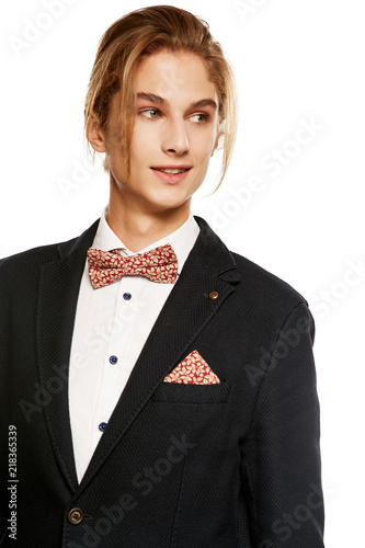 A young man in a black suit jacket and button up shirt, accessorized with a red fleaf pattern  bow tie and matching pocket square. The blond smiling while looking aside over the white background.  photo